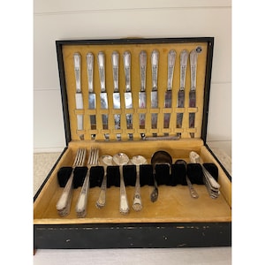 May include: A silver-toned flatware set arranged in a black wooden storage case with a tan velvet interior. The set includes knives with ornate handles, forks, and various spoons, all featuring a consistent decorative pattern on the metal handles.