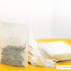 May include: Close-up of several white tea bags with attached strings, filled with loose tea leaves. The tea bags are stacked on a yellow surface, with one bag standing upright, showcasing the tea inside. The image is well-lit, highlighting the tea's texture.