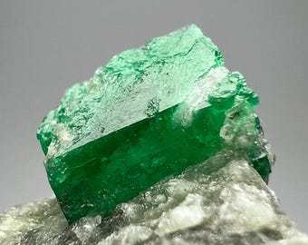Puede incluir: Primer plano de un cristal verde esmeralda vibrante, de forma rectangular, sobre una base mineral clara y texturizada. La superficie del cristal brilla, revelando su claridad y color intenso. La imagen resalta la belleza natural de la piedra preciosa.