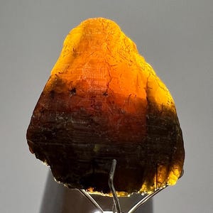 Peut inclure: Une tranche de pierre précieuse translucide de couleur ambre, avec un dégradé allant du marron foncé à l'orange vif. La surface brute est éclairée, révélant des textures et des motifs naturels. La pierre est montée sur un support en métal argenté.
