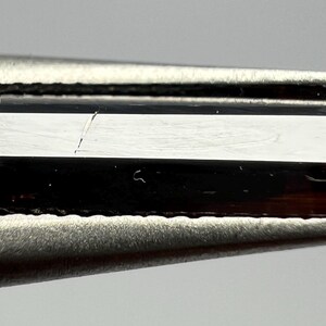 Puede incluir: Una gema rectangular facetada, de color marrón oscuro, sujeta con unas pinzas. La gema tiene una superficie reflectante más clara con una pequeña grieta. Los bordes están bien definidos. Las pinzas son de color plateado.