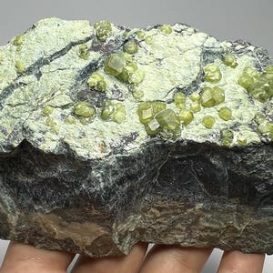 Könnte beinhalten: Eine grob behauene Felsformation mit einer dunkelgrauen Basis und einer hellgrünen, strukturierten Oberseite. Zahlreiche kleine, hellgrüne Kristalle sind über die Oberfläche verstreut. Der Felsen wird in der Hand einer Person gehalten.
