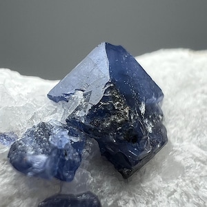 May include: Close-up of a cluster of blue crystals with sharp, geometric shapes. The crystals have a deep indigo color with some areas appearing nearly black. They are set against a backdrop of white, crystalline material.