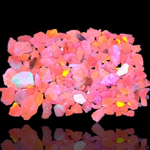 May include: A collection of small, irregularly shaped pink and orange rocks, possibly gemstones or minerals, against a black background. Some rocks have hints of yellow and green. The image has a reflective surface.