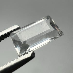 May include: A clear, rectangular gemstone with multiple facets is held by a pair of silver tweezers. The gemstone is transparent, allowing light to pass through, and has a polished appearance. The tweezers are in focus, highlighting the gem's clarity.