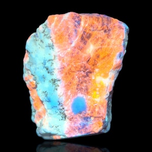 May include: A vibrant, irregularly shaped rock with a striking color palette. The stone features a mix of orange, blue, and purple hues, with a glossy finish. The image is set against a black background, highlighting the rock's unique texture and color variations.