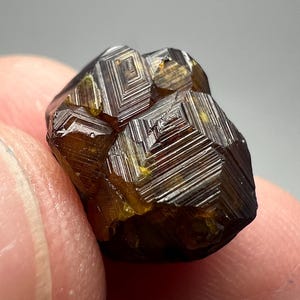 May include: A close-up of a faceted, dark amber-colored mineral specimen. The crystal has a geometric pattern of lines and planes, with a slightly rough texture. The mineral is held between two fingers.