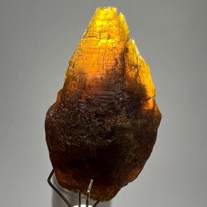 Könnte beinhalten: Ein roher, ungeschliffener Edelstein in einem satten Bernsteinton, der an der Basis in ein dunkleres Braun übergeht. Die Oberfläche hat ein raues, strukturiertes Aussehen mit sichtbaren kristallinen Strukturen. Der Edelstein ist auf einem Metallständer montiert.