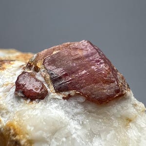 Puede incluir: Primer plano de una gema en bruto sin cortar. La gema es de un color marrón rojizo intenso con una estructura cristalina. Se encuentra sobre una base mineral blanca y beige, mostrando su forma y textura naturales.