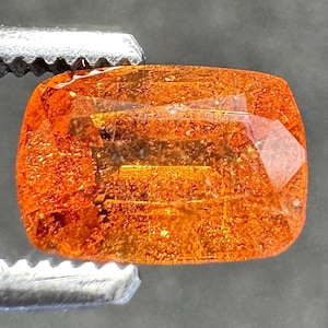 May include: Close-up of a vibrant, orange gemstone with a rectangular shape and rounded corners. The gem is held by a silver tweezer, showcasing its internal structure and reflective surfaces. The gemstone's color ranges from amber to a deep orange.