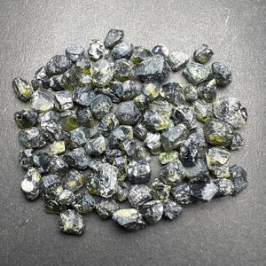 May include: A collection of raw, uncut gemstones. The stones are a mix of dark gray and olive green, with a faceted, crystalline appearance. The gemstones are scattered across a dark surface, showcasing their natural textures and colors.