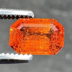 May include: A faceted, rectangular orange gemstone held by a metal tweezer. The gem has a vibrant, translucent appearance with multiple facets reflecting light. The gemstone's color ranges from a deep orange to a lighter, almost yellow hue.
