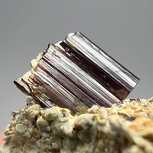 Puede incluir: Primer plano de un espécimen mineral con un grupo de cristales metálicos de color marrón rojizo. Los cristales tienen una apariencia estriada y están colocados sobre una matriz beige rugosa. La iluminación resalta las superficies reflectantes de los cristales.