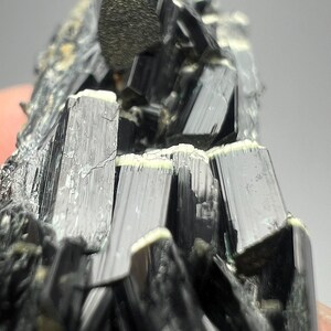 Puede incluir: Primer plano de un grupo de cristales de turmalina negra. Los cristales son alargados y tienen una superficie brillante y reflectante. Algunos cristales tienen un borde de color claro. La apariencia general es oscura y texturizada, con ángulos agudos y una estética natural y cruda.