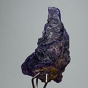 May include: A raw, natural amethyst crystal specimen. The deep purple and black stone has a rough, textured surface and is mounted on a metal stand. The crystal's jagged edges and varying shades of color create a striking visual.