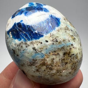 Può includere: Una pietra levigata di forma ovale caratterizzata da un mix vibrante di blu reale intenso, bianco brillante e sezioni grigio pallido. La superficie presenta una texture cristallina naturale con macchie minerali marrone scuro e nere sparse.
