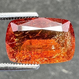May include: A faceted, rectangular gemstone with a warm orange and reddish-brown color. The gem is held by a silver tweezer, showcasing its polished surfaces and internal inclusions. The gem's shape is a modified cushion cut.