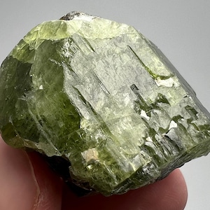 May include: A rough-cut, olive-green peridot gemstone. The crystal has a faceted, irregular shape with a glossy, reflective surface. The stone is held in a hand, showcasing its natural, unpolished beauty.