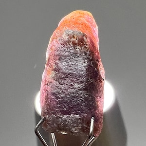 Op de afbeelding: Een ruwe, ongeslepen edelsteen met een kleurengradiënt. De bovenkant is rood-oranje, overgaand in een diep paars aan de basis. De steen wordt vastgehouden door metalen klemmen, die de natuurlijke, onbewerkte textuur laten zien.