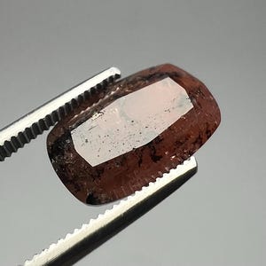 以下が含まれることがあります： 銀色のピンセットで保持された、ファセット加工された赤褐色の宝石。 宝石は、丸みを帯びた角と反射面を持つ長方形です。 石には暗いインクルージョンと明るい色の内部があります。