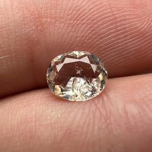 Pode incluir: Uma gema facetada em forma oval com um tom amarelo pálido. A gema tem múltiplas facetas que refletem a luz, criando um efeito brilhante. A pedra é segurada na mão de uma pessoa, com a pele visível ao fundo.