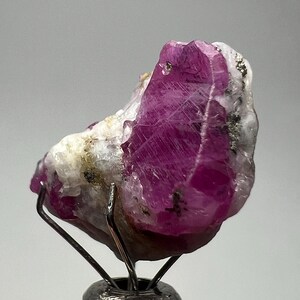 May include: A raw ruby gemstone with a deep magenta hue, set on a metal stand. The ruby is partially encased in white and gray matrix, with visible crystalline structures. Small flecks of other minerals are present.