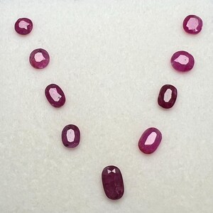 Puede incluir: Una colección de diez rubíes facetados, de formas ovaladas y redondas. Las piedras preciosas son de un color rojo intenso y se disponen sobre una superficie blanca. Los rubíes varían en tamaño, algunos parecen más alargados que otros. La disposición destaca las gemas.