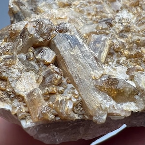 May include: Close-up of a natural mineral specimen featuring clusters of translucent, light brown crystals. The crystals have a prismatic shape and are set against a rough, beige matrix. The overall texture is rough and crystalline.