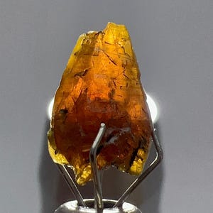 May include: A translucent, amber-colored gemstone with a rough, irregular shape. The stone is held by a silver metal stand, and it has dark inclusions throughout. The lighting highlights the gem's internal structure and color variations.