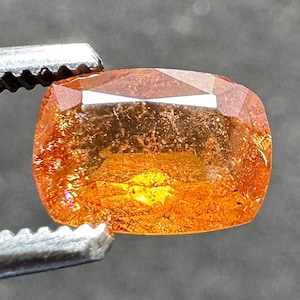 May include: A faceted, orange-yellow gemstone held by a metal tweezer. The gem has a rectangular cushion cut with visible internal inclusions and a bright, reflective surface. The gem's color ranges from a deep orange to a lighter yellow.