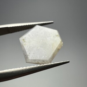 May include: A clear, hexagonal gemstone held by a pair of tweezers. The gem has a translucent quality with subtle variations in color and texture. The tweezers are silver and the background is a soft gray.