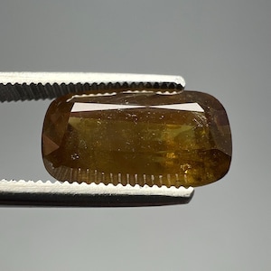 Puede incluir: Una gema facetada, rectangular con esquinas redondeadas, sostenida por unas pinzas plateadas. La gema tiene un color marrón dorado cálido con reflejos internos. Las pinzas están enfocadas, resaltando la claridad y el corte de la gema.