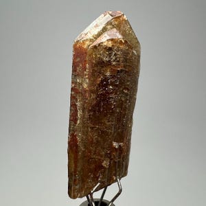 May include: A natural, raw mineral specimen. The crystal is a warm amber color with reddish-brown inclusions and a textured surface. The mineral is mounted on a silver-colored stand, showcasing its natural form and color variations.