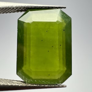 May include: A green gemstone with a rectangular emerald cut. The gemstone has a few small black inclusions.