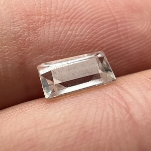 May include: A clear, rectangular gemstone with a faceted cut. The gem is held between fingers, showcasing its clarity and shape. The gemstone appears to be colorless, reflecting light.