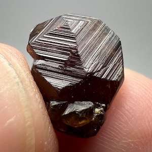 May include: Close-up of a dark brown, faceted mineral specimen. The crystal has a geometric shape with a series of parallel lines across its surface. The mineral is held between two fingers.
