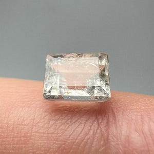 Pode incluir: Uma gema clara, retangular, com um corte facetado. A gema é segurada entre os dedos, mostrando sua clareza e forma. A pedra parece ser um cristal natural, refletindo a luz e exibindo características internas.