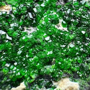 Pode incluir: Close-up de um aglomerado de cristais verde-esmeralda vibrantes. Os cristais têm uma estrutura geométrica facetada, refletindo a luz e criando um efeito cintilante. Os cristais estão sobre um fundo escuro e texturizado.