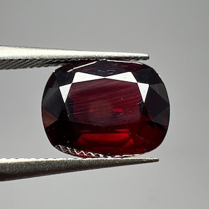 May include: A faceted, oval-shaped, deep red gemstone held by silver tweezers. The gem has a polished surface with multiple facets that reflect light, creating a sparkling effect. The gemstone's rich color and clarity are highlighted against a neutral background.