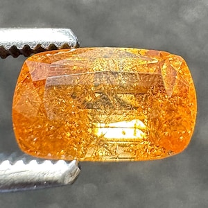 May include: A faceted, orange-yellow gemstone held by a silver tweezer. The gem has a rectangular shape with rounded corners and internal inclusions. The stone's color ranges from a deep amber to a lighter, golden hue, with visible internal structures.