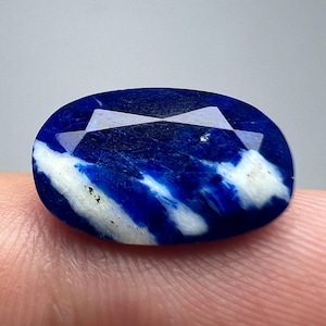 May include: Oval-shaped gemstone with a deep blue hue, accented by white streaks. The gem features a faceted top, creating a play of light. The stone is a striking example of natural mineral beauty.