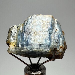 May include: A natural kyanite crystal, showing a layered structure with blue and white hues, and a hint of gold. The crystal is mounted on a metal stand, highlighting its natural form and texture. A mineral specimen.