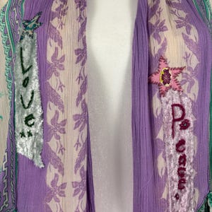 May include: A draped, open-front garment in shades of purple, cream, and green. The fabric features a floral pattern and is embellished with appliqued patches that spell out "Love" and "Peace". The garment has a bohemian aesthetic.