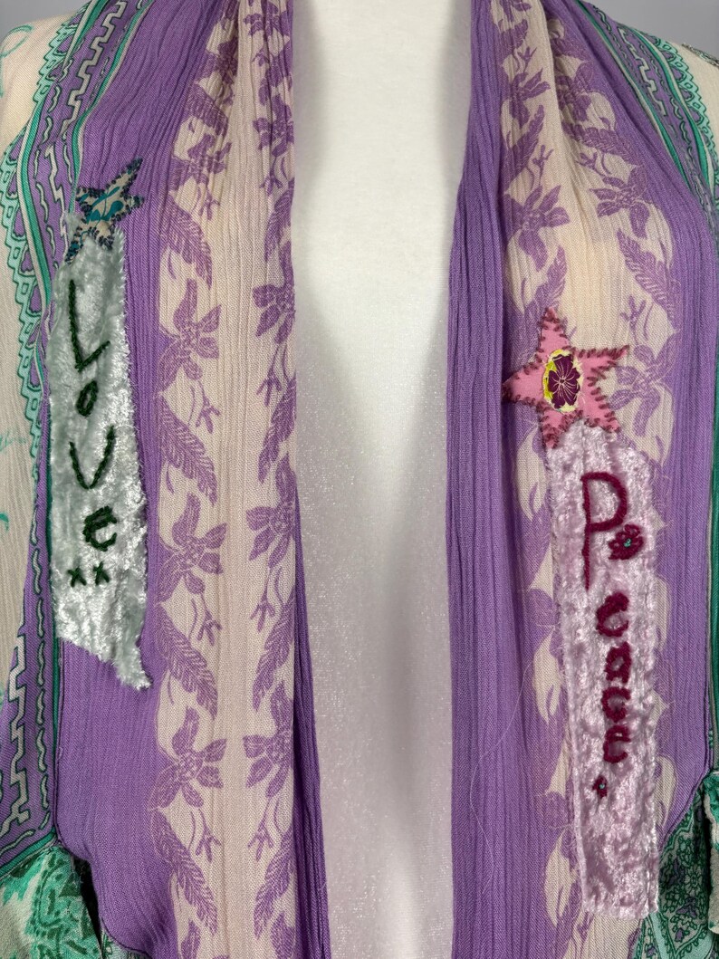 May include: A draped, open-front garment in shades of purple, cream, and green. The fabric features a floral pattern and is embellished with appliqued patches that spell out "Love" and "Peace". The garment has a bohemian aesthetic.