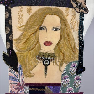 May include: A fabric art piece featuring a portrait of a woman with long blonde hair, red lips, and a beaded necklace. The artwork is framed with patterned fabric and embellished with decorative elements, including a butterfly and star.