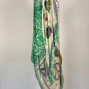 May include: A flowing, patterned garment in shades of green, cream, and purple. The design includes paisley, floral, and other decorative motifs. The garment is draped over a white mannequin, showcasing its shape and details.