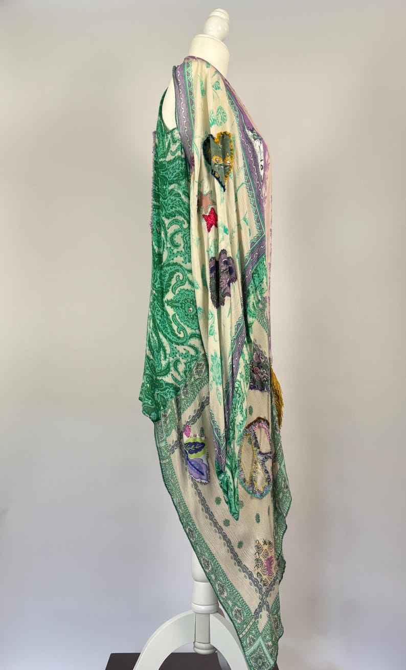 May include: A flowing, patterned garment in shades of green, cream, and purple. The design includes paisley, floral, and other decorative motifs. The garment is draped over a white mannequin, showcasing its shape and details.
