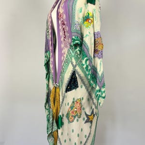 May include: A cream-colored kimono with a patchwork design featuring floral and geometric patterns in shades of green, purple, and yellow. The kimono has a long, flowing silhouette and is adorned with tassels and embellishments.