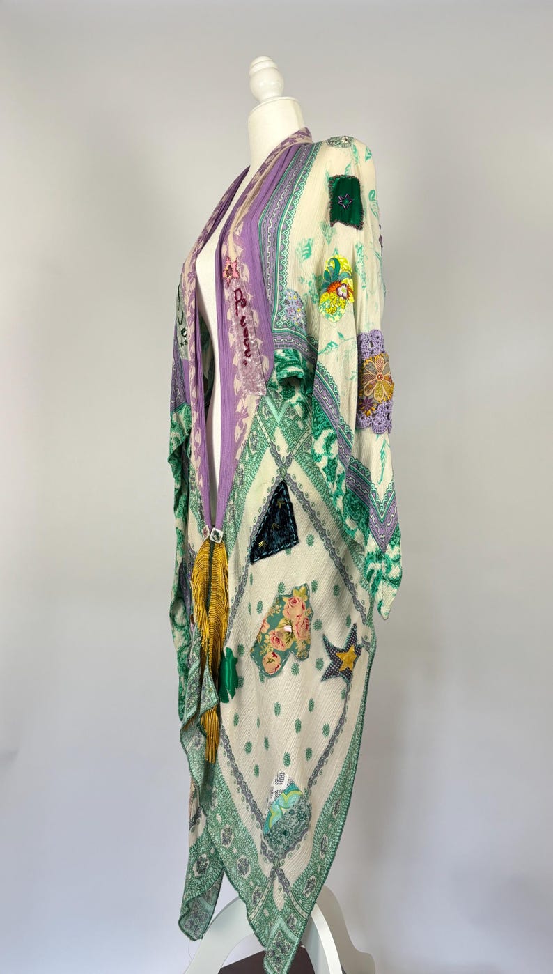May include: A cream-colored kimono with a patchwork design featuring floral and geometric patterns in shades of green, purple, and yellow. The kimono has a long, flowing silhouette and is adorned with tassels and embellishments.
