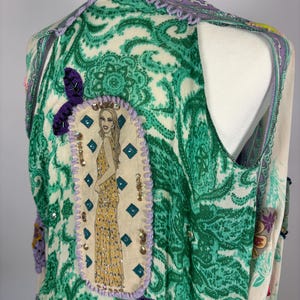May include: A sleeveless vest with a paisley pattern in green and cream. The vest features a purple trim and a decorative patch with a woman in a yellow dress. The vest is adorned with sequins and purple leaf accents.